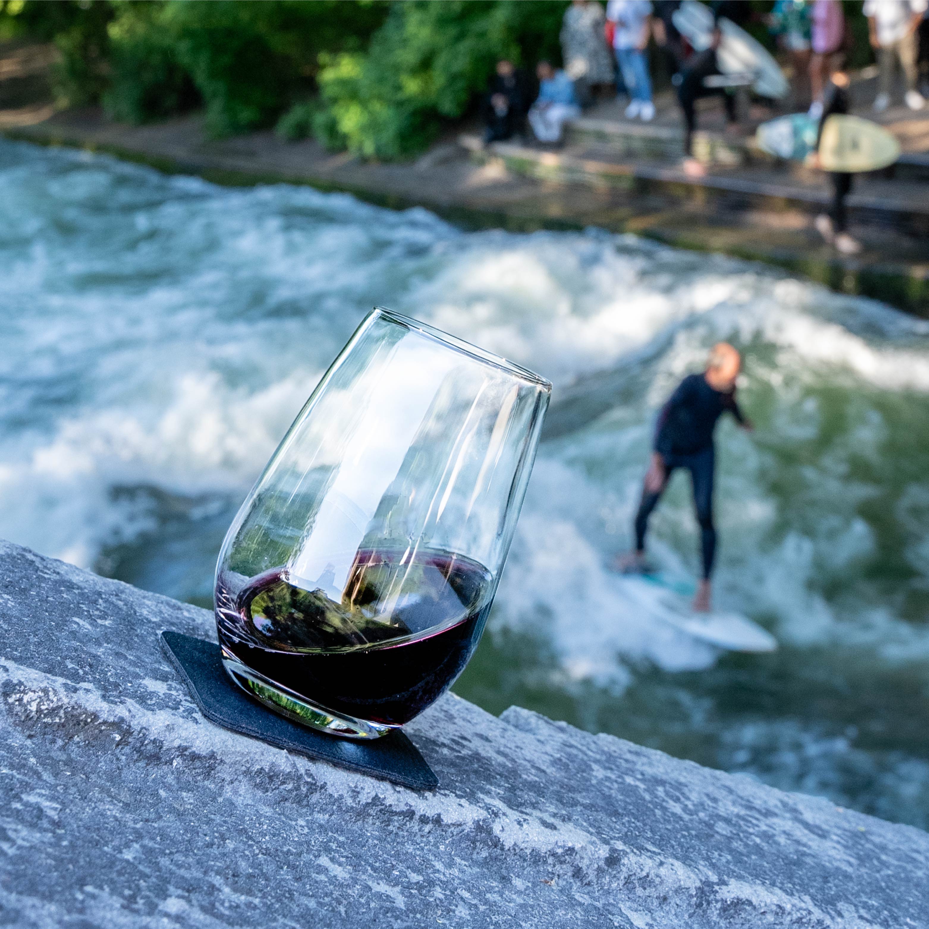 Magnet-Kristallglas Longdrink auf quadratischem Metall-Nano-Gel-Pad vor Eisbachwelle in München mit Surfer im Hintergrund 